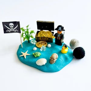 Peut inclure: Un ensemble de jeu sur le thème des pirates sur une surface bleue. Il comprend un drapeau pirate, un coffre au trésor avec des pièces d'or, une figurine de pirate Lego et divers objets miniatures sur le thème de la mer. Deux boules de feutre et un canard en caoutchouc complètent la scène.