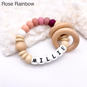 May include: A wooden teething ring with a silicone bead chain. The beads are in shades of pink, beige, and white. The ring has a name spelled out in white silicone beads: "MILLIE".
