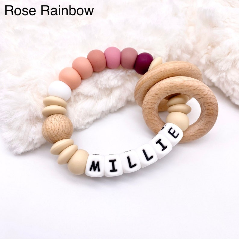 May include: A wooden teething ring with a silicone bead chain. The beads are in shades of pink, beige, and white. The ring has a name spelled out in white silicone beads: "MILLIE".