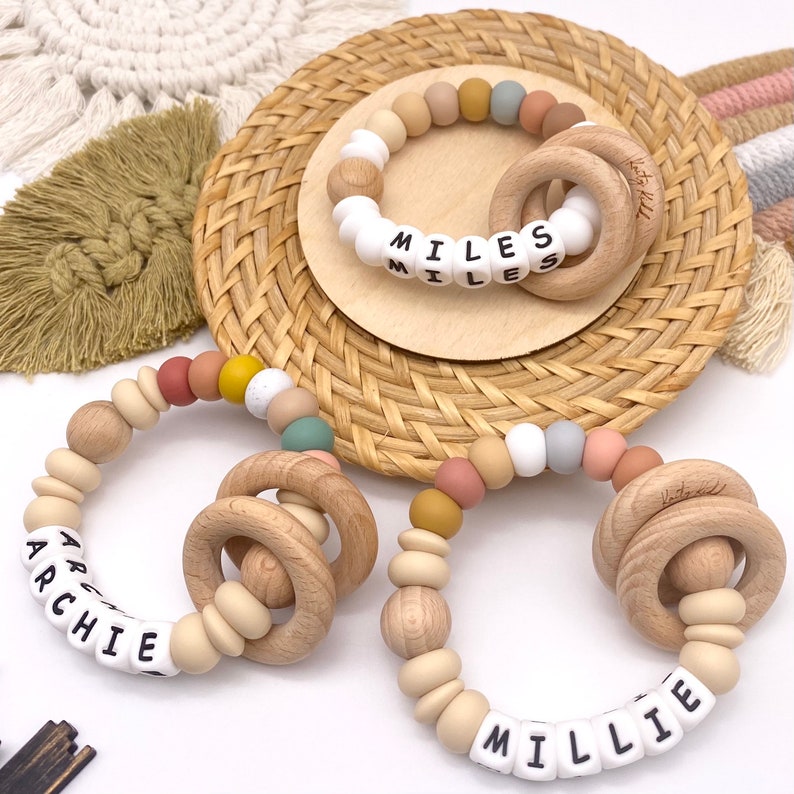 May include: Two wooden teething rings with silicone beads and personalized name tags. The rings are in natural wood with a smooth finish. The beads are in various colors, including beige, light brown, pink, yellow, and gray. The name tags are white with black lettering. The names are "Archie" and "Millie".