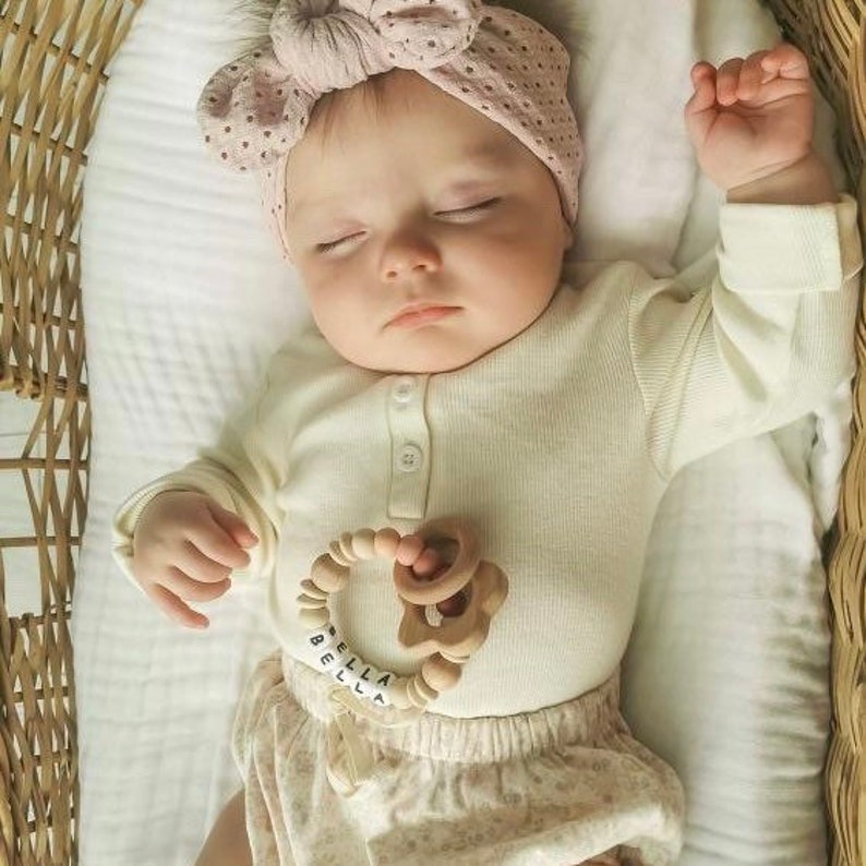 May include: A baby wearing a white onesie with a pink polka dot headband and a wooden teething toy with the name "BELLA" spelled out in black letters.