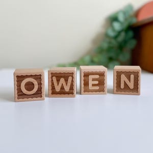 May include: Four wooden blocks spelling "OWEN". Each block is light brown with a darker brown letter. The blocks have a decorative scalloped edge. The blocks are arranged in a row on a white surface.