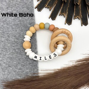 May include: A white and natural wood baby teether with the name "MILES" spelled out in white beads. The teether has a ring of colorful silicone beads and a wooden ring.