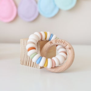 May include: A wooden teething ring with the name "Bobby" engraved on it. The ring is decorated with a colorful silicone bead bracelet.