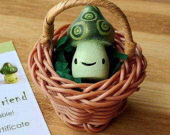 May include: A small green and white mushroom figurine with a smiling face sits in a brown wicker basket lined with green shredded paper.