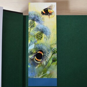 May include: A bookmark with a watercolor illustration of two bumblebees on a thistle plant. The bookmark has a blue border at the bottom.