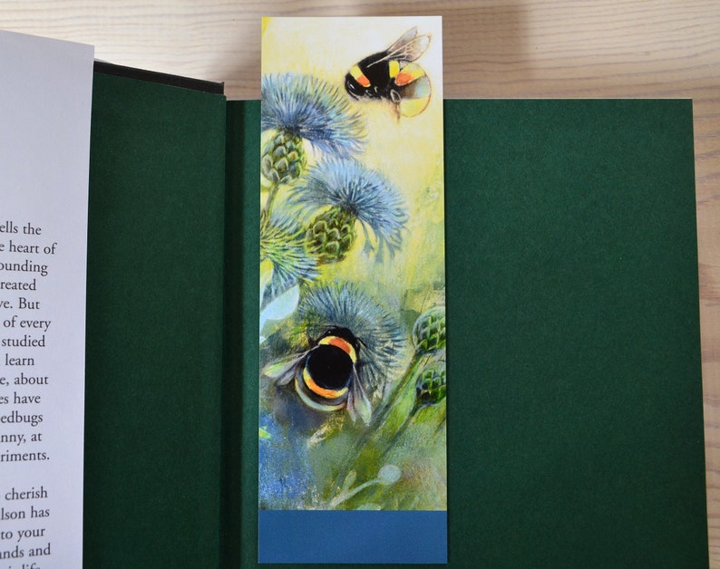 May include: A bookmark with a watercolor illustration of two bumblebees on a thistle plant. The bookmark has a blue border at the bottom.