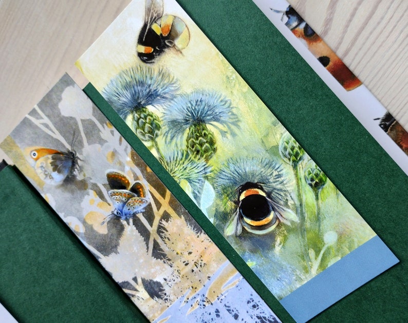 May include: Three bookmarks with watercolor illustrations of insects and flowers. The first bookmark features a blue butterfly on a light gray background. The second bookmark features a bumblebee on a blue and green background. The third bookmark features a bumblebee on a yellow and green background.