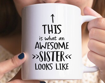 May include: White ceramic mug with black text that reads "This is what an awesome sister looks like" with an arrow pointing up.