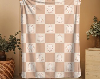 May include: A beige and white checkered blanket featuring a repeating pattern of cartoon ghosts. The ghosts are white with peach-colored details. The blanket is held up by a person.