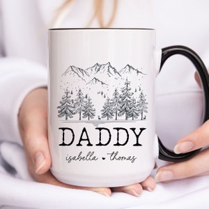 May include: White ceramic mug with a black handle. The mug features a black and white illustration of a mountain range with trees and the text "DADDY isabella thomas".