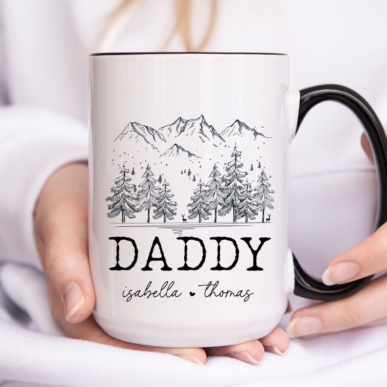 May include: White ceramic mug with a black handle. The mug features a black and white illustration of a mountain range with trees and the text "DADDY isabella thomas".