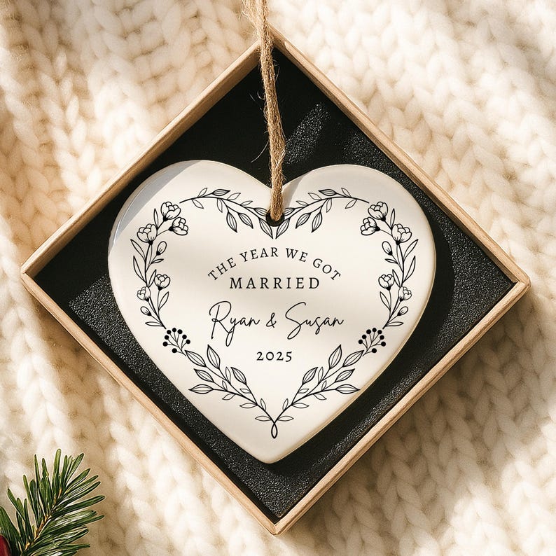 K&ouml;nnte beinhalten: Herzf&ouml;rmiger Keramikschmuck mit floralem Kranzdesign. Der Schmuck tr&auml;gt den Text "THE YEAR WE GOT MARRIED" &uuml;ber den Namen "Ryan & Susan" und dem Jahr "2025". Pr&auml;sentiert in einer Geschenkbox mit einer Schnur zum Aufh&auml;ngen.