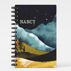 May include: A black and blue watercolor notebook with a mountain landscape and a starry night sky. The notebook has a black spiral binding and the name "NANCY" printed on the front cover.