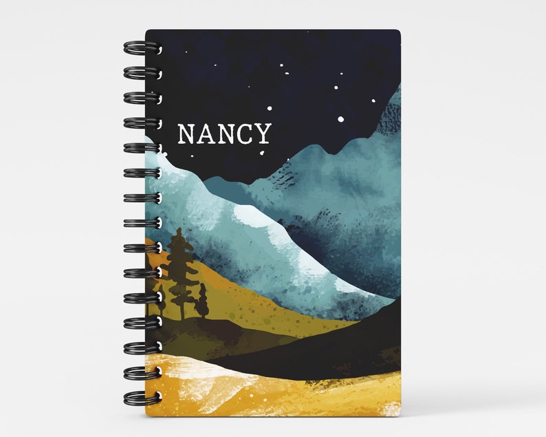 May include: A black and blue watercolor notebook with a mountain landscape and a starry night sky. The notebook has a black spiral binding and the name "NANCY" printed on the front cover.