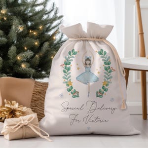 May include: White canvas gift bag with a drawstring closure. The bag features a watercolor illustration of a ballerina in a blue tutu and a green wreath with gold stars. The text "Special Delivery for Victoria" is printed on the bag.