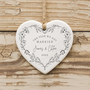 K&ouml;nnte beinhalten: Wei&szlig;es herzf&ouml;rmiges Ornament mit floralem Kranzdesign und Juteschnur zum Aufh&auml;ngen. Der Text "THE YEAR WE GOT MARRIED James & Chloe 2025" ist in schwarzer Schrift dargestellt. Das Ornament liegt auf Holz.