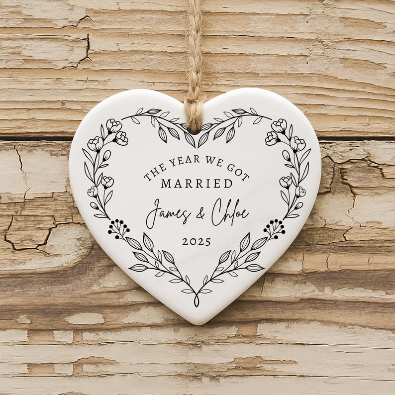 K&ouml;nnte beinhalten: Wei&szlig;es herzf&ouml;rmiges Ornament mit floralem Kranzdesign und Juteschnur zum Aufh&auml;ngen. Der Text "THE YEAR WE GOT MARRIED James & Chloe 2025" ist in schwarzer Schrift dargestellt. Das Ornament liegt auf Holz.