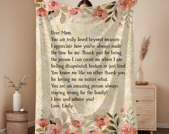 Pode incluir: Um cobertor creme com detalhes florais e o texto "Dear Mom, You are truly loved beyond measure." O cobertor apresenta um design floral com cores pêssego e verde. O cobertor é um item de decoração aconchegante.