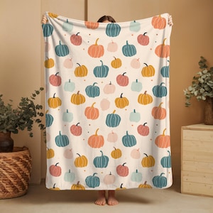 Può includere: Una coperta color crema con un motivo ripetuto di zucche colorate nei toni dell'arancione, verde acqua, rosa e giallo. Le zucche hanno steli neri e sono sparse sulla superficie. La coperta è tenuta da una persona.
