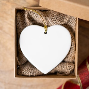 K&ouml;nnte beinhalten: Ein wei&szlig;er herzf&ouml;rmiger Schmuck mit goldenem Faden, pr&auml;sentiert in einer braunen Geschenkbox mit Jutefutter. Ein rot-goldenes Band ist sichtbar. Der Schmuck ist leer und bereit zur Personalisierung.