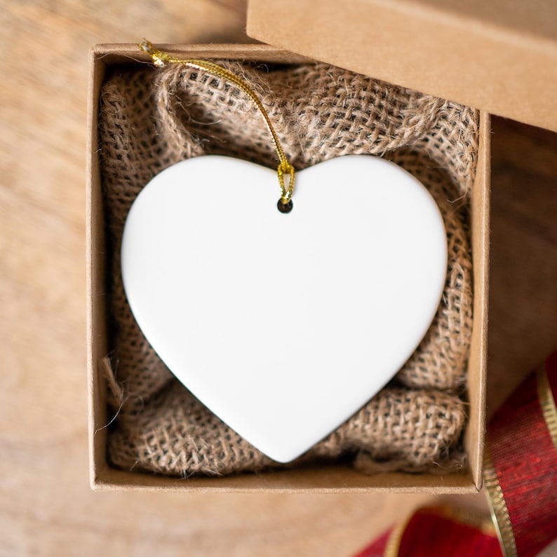 K&ouml;nnte beinhalten: Ein wei&szlig;er herzf&ouml;rmiger Schmuck mit goldenem Faden, pr&auml;sentiert in einer braunen Geschenkbox mit Jutefutter. Ein rot-goldenes Band ist sichtbar. Der Schmuck ist leer und bereit zur Personalisierung.