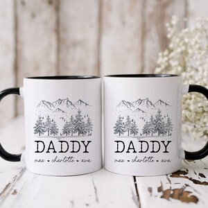 May include: Two white ceramic mugs with black rims and handles. Each mug features a black and white illustration of a mountain range with pine trees and the word "Daddy" with names below.