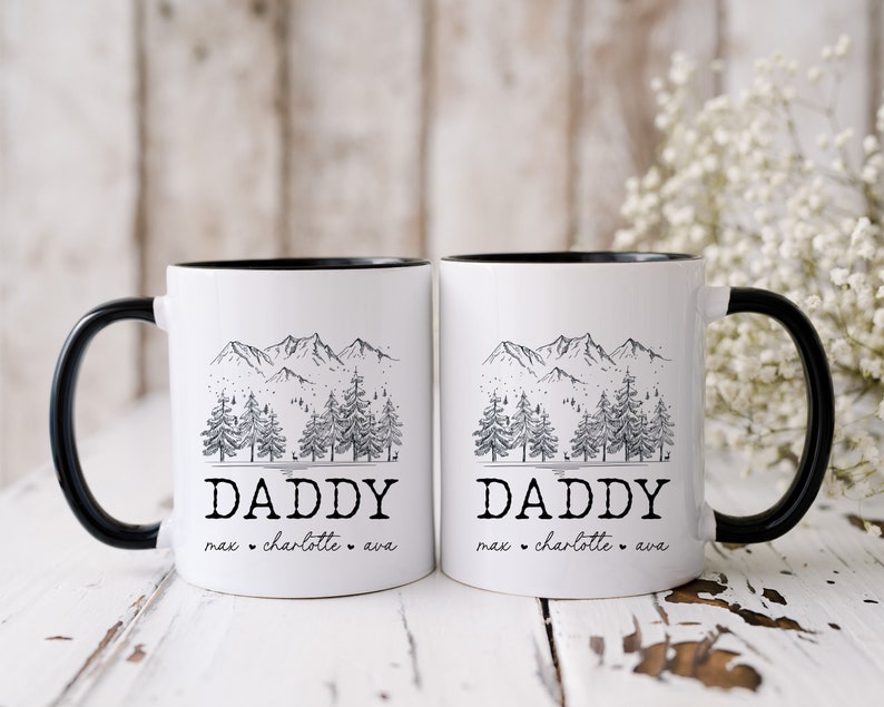 May include: Two white ceramic mugs with black rims and handles. Each mug features a black and white illustration of a mountain range with pine trees and the word "Daddy" with names below.
