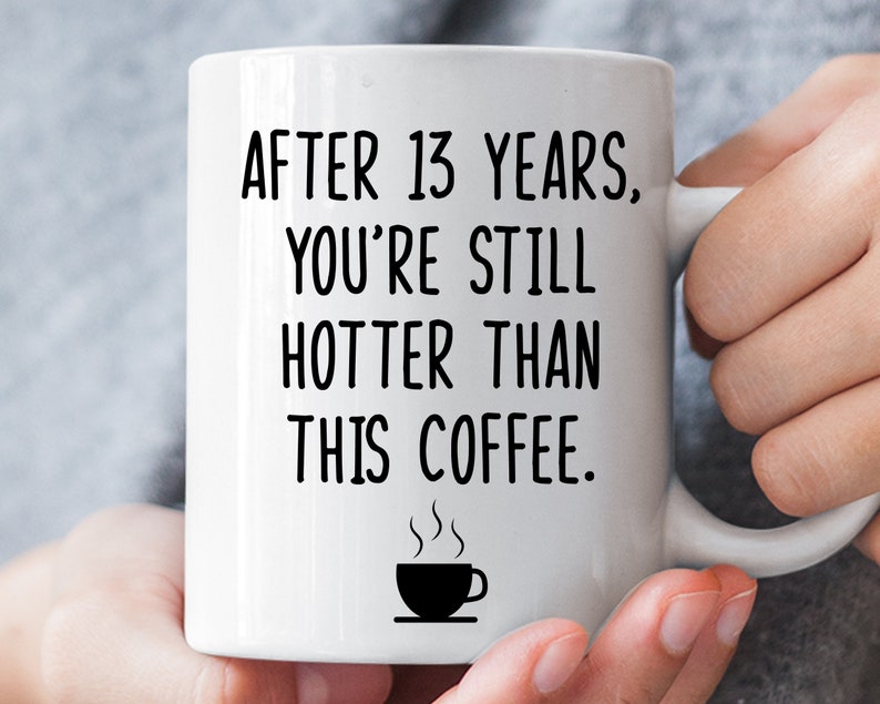 May include: White ceramic coffee mug with black text that reads "After 13 years, you're still hotter than this coffee."  A small black coffee cup with steam is below the text.