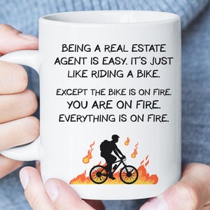 May include: White ceramic coffee mug with a black silhouette of a person riding a bicycle on fire. The text on the mug reads "Being a real estate agent is easy. It's just like riding a bike. Except the bike is on fire. You are on fire. Everything is on fire."