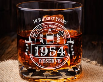 May include: A clear glass whiskey tumbler with a white design that reads "In Whiskey Years You Just Got More Delicious 1954 Reserve". The glass is filled with amber liquid and ice cubes.