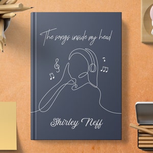 Könnte beinhalten: Ein dunkelblaues Notizbuch mit dem Text "The songs inside my head" und dem Namen "Shirley Neff" in weißer Kursivschrift. Eine Strichzeichnung einer Person mit Kopfhörern und Musiknoten um sie herum.