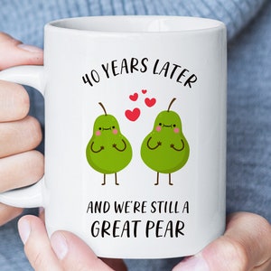 Op de afbeelding: Witte keramische mok met een illustratie van een groen paar peren en de tekst "40 jaar later en we zijn nog steeds een geweldige peer".
