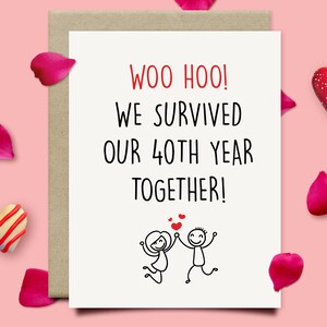 May include: A greeting card with the text "WOO HOO! WE SURVIVED OUR 40TH YEAR TOGETHER!" in black and red. The card features a stick figure couple holding hands. Pink rose petals and heart-shaped decorations surround the card.