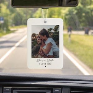 May include: A rectangular car air freshener with a photo of a couple hugging. The air freshener has the words "Drive Safe" and "I LOVE YOU" printed on it, with a small heart symbol. It hangs from a white string.