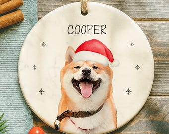 May include: A round ceramic Christmas ornament featuring a smiling dog wearing a Santa hat. The dog's name, "COOPER", is printed above the dog. The ornament has a string for hanging and snowflake designs.