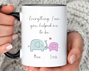 May include: White ceramic mug with a black rim and handle. The mug features a hand-drawn illustration of two elephants holding trunks, with the text "Everything I am you helped me to be" and the names "Mom" and "Lisa" below.