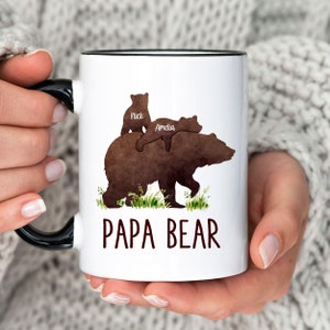 K&ouml;nnte beinhalten: Eine wei&szlig;e Keramiktasse mit schwarzem Rand. Die Tasse zeigt eine braune Aquarell-Illustration eines B&auml;ren mit zwei kleineren B&auml;ren auf seinem R&uuml;cken. Der Text "Nick" und "Amelia" steht auf den kleineren B&auml;ren. Der Text "Papa Bear" steht unter den B&auml;ren.