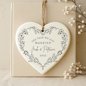K&ouml;nnte beinhalten: Herzf&ouml;rmiger Keramikschmuck mit floralem Kranzdesign und dem Text "THE YEAR WE GOT MARRIED Jack & Patricia 2025". Der Schmuck h&auml;ngt an einer Schnur aus Naturfaser. Der Hintergrund ist neutral gef&auml;rbt.