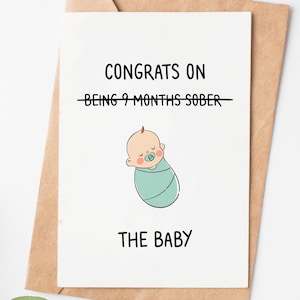 May include: A white card with black text that reads "Congrats on being 9 months sober" with a line through "sober" and the word "the baby" below. A cartoon illustration of a baby swaddled in a blue blanket is above the text.