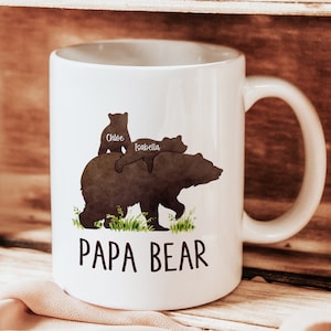 K&ouml;nnte beinhalten: Wei&szlig;e Keramiktasse mit einer Silhouette einer B&auml;renfamilie. Der Vaterb&auml;r geht mit zwei Jungen auf dem R&uuml;cken. Die Jungen sind mit "Chloe" und "Isabella" beschriftet. Die Tasse tr&auml;gt die Aufschrift "Papa Bear".