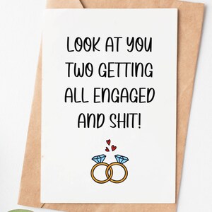 May include: A white card with black text that reads "Look at you two getting all engaged and shit!" with a cartoon drawing of two gold rings with blue diamonds and red hearts above them.