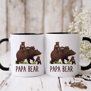 K&ouml;nnte beinhalten: Zwei wei&szlig;e Keramikbecher mit schwarzen R&auml;ndern und Griffen. Jeder Becher zeigt eine braune B&auml;rensilhouette mit drei kleineren B&auml;rensilhouetten oben, die eine Familie darstellen. Der Text "Papa Bear" ist auf der Vorderseite jedes Bechers aufgedruckt.