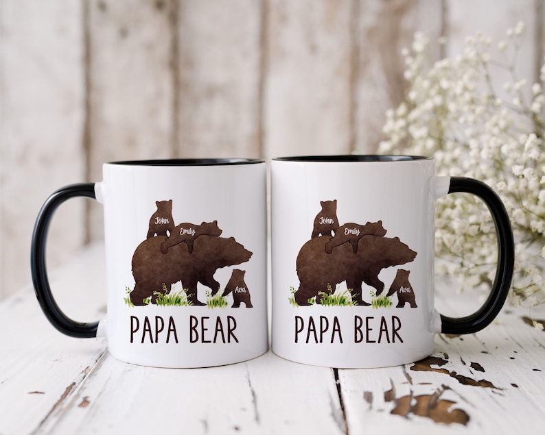 K&ouml;nnte beinhalten: Zwei wei&szlig;e Keramikbecher mit schwarzen R&auml;ndern und Griffen. Jeder Becher zeigt eine braune B&auml;rensilhouette mit drei kleineren B&auml;rensilhouetten oben, die eine Familie darstellen. Der Text "Papa Bear" ist auf der Vorderseite jedes Bechers aufgedruckt.