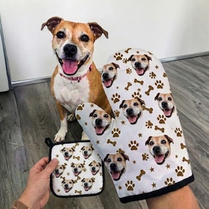 May include: A smiling brown and white dog sits next to a white oven mitt and pot holder. The mitt and pot holder are decorated with images of the dog's face, paw prints, and bones.