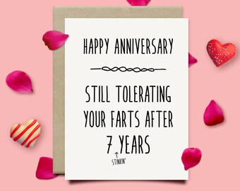 May include: A white card with black text that reads "Happy Anniversary - Still Tolerating Your Farts After 7 Years - Stinkin'"  The card is surrounded by pink rose petals and red hearts.