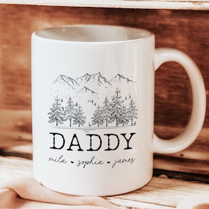 May include: White ceramic mug with a black and white illustration of a mountain range and pine trees. The text "DADDY" is printed below the illustration, with the names "mila", "sophia", and "james" printed below the text.