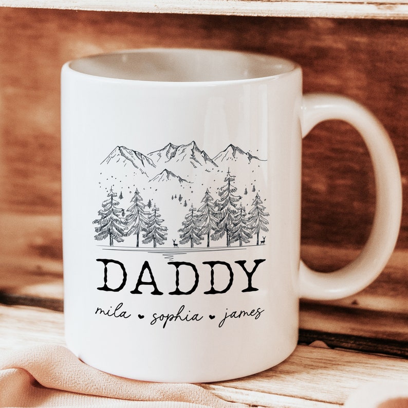 May include: White ceramic mug with a black and white illustration of a mountain range and pine trees. The text "DADDY" is printed below the illustration, with the names "mila", "sophia", and "james" printed below the text.