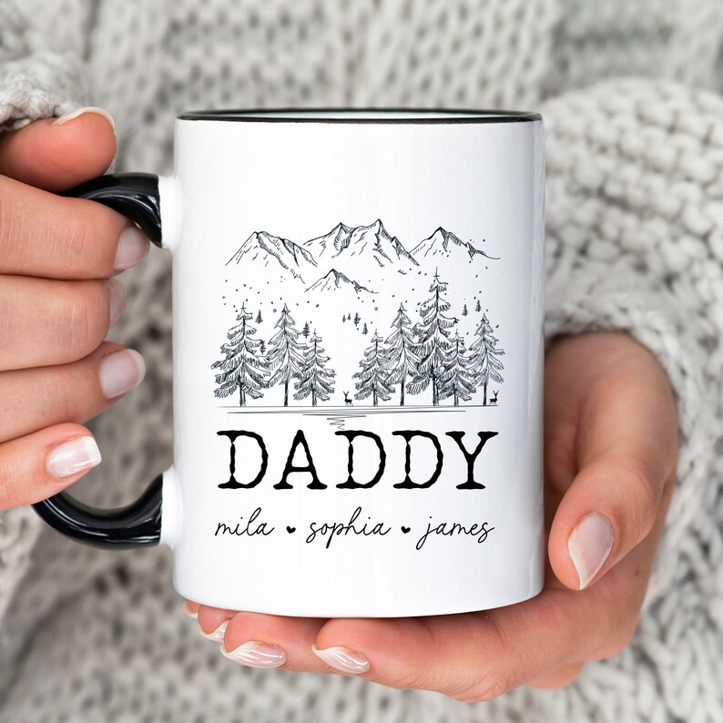 May include: White ceramic mug with a black rim and handle. The mug features a black and white illustration of a mountain range with pine trees and the word "DADDY" below. The names "mila", "sophia", and "james" are written below the word "DADDY".
