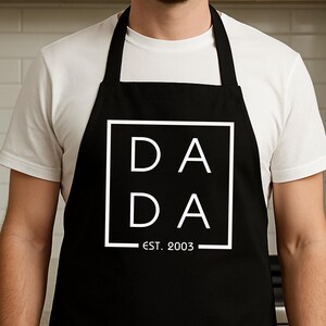 以下が含まれることがあります： 白い正方形の中に「DADA」という文字が入った黒いエプロン。その下に「EST. 2003」と書かれています。白いTシャツの上からエプロンを着用しています。